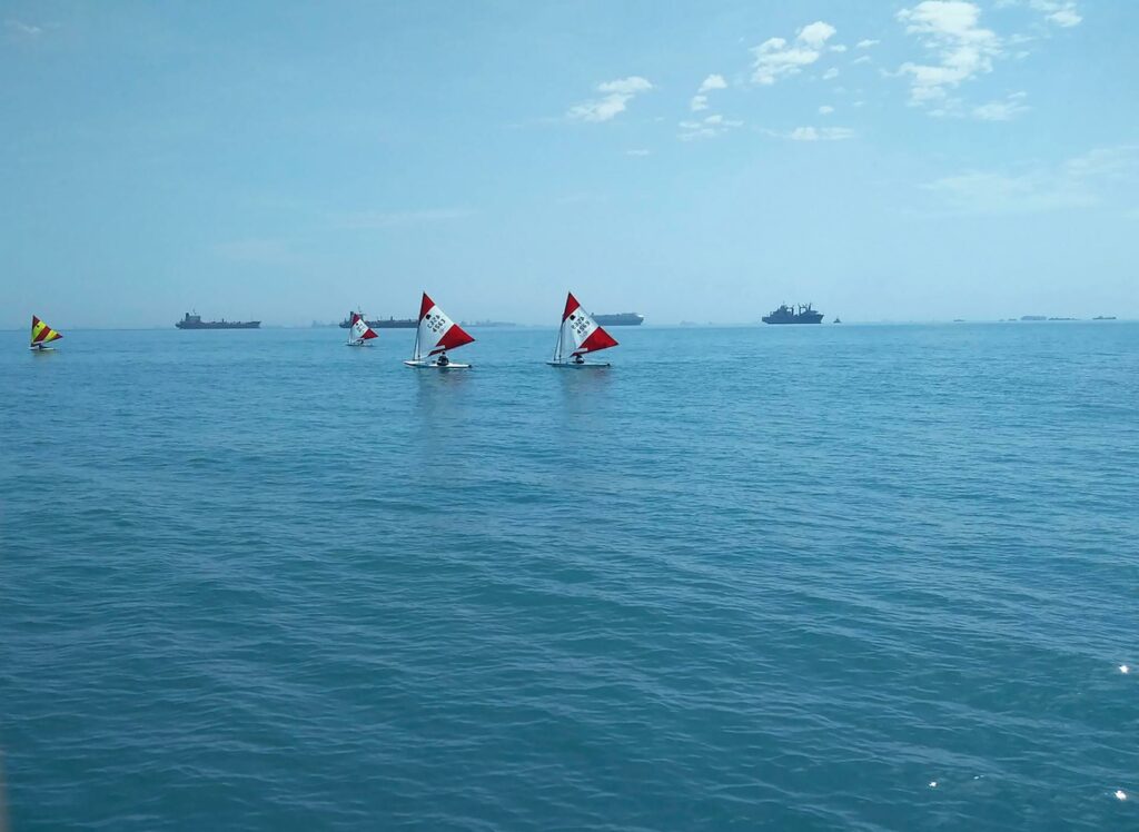 Several small sailboats with red and white sails glide on calm blue water under a mostly clear sky, while larger ships are visible on the distant horizon.