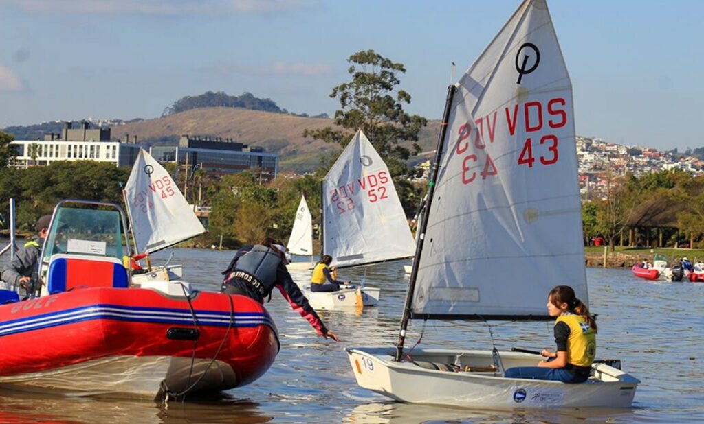 Children in small sailboats participate in a sailing activity on a lake, supervised by an adult on a red motorboat. Trees, buildings, and hills are visible in the background under a blue sky.