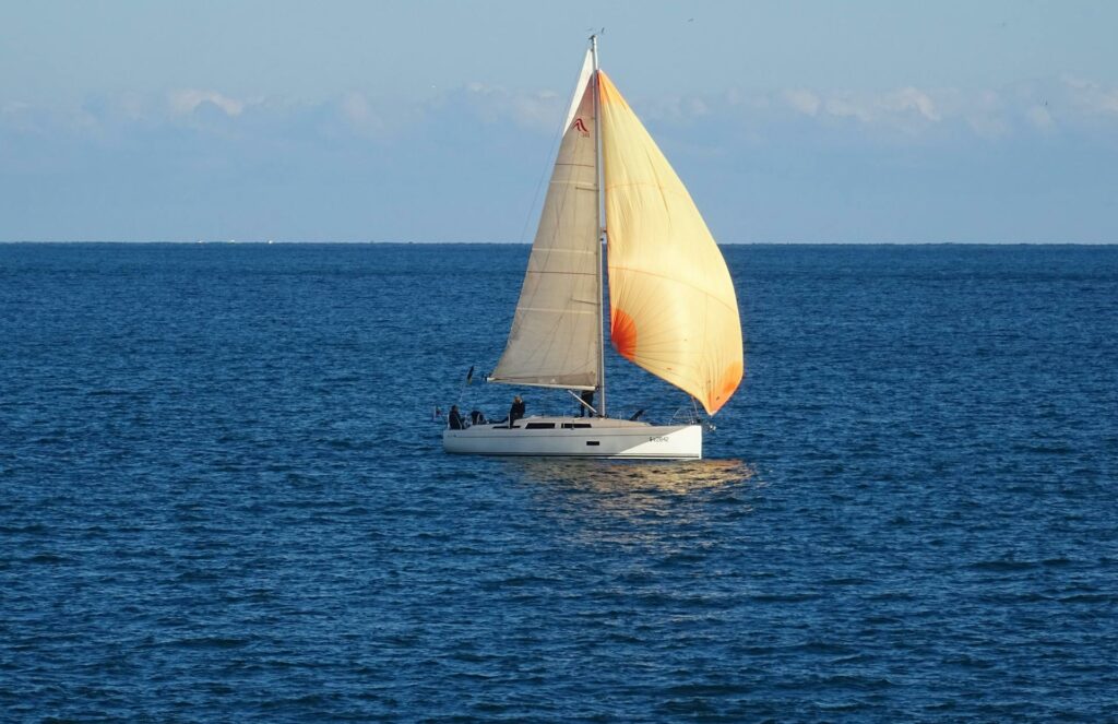 A white sailboat with tan sails glides across a calm, blue sea under a clear sky, with a few people visible on board.