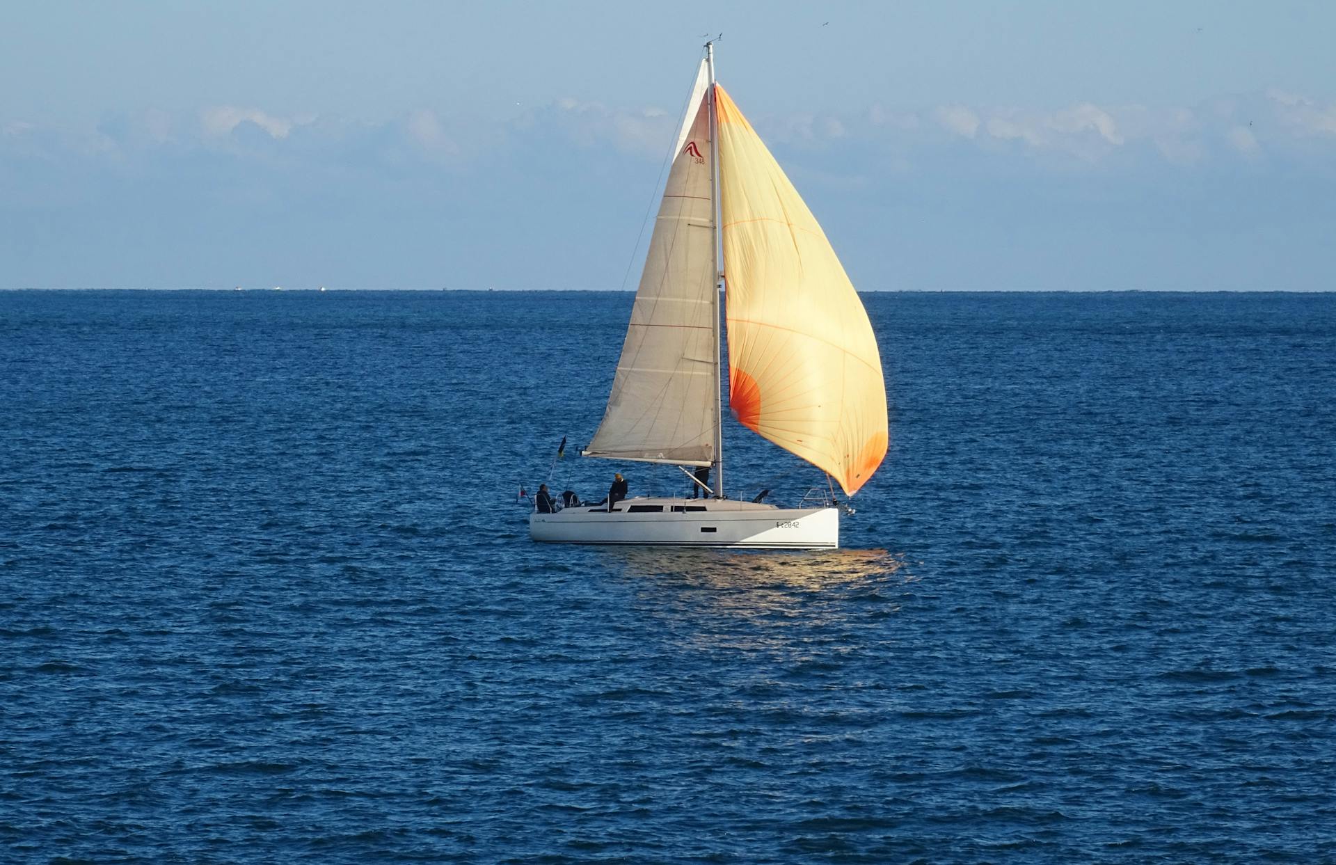 A white sailboat with tan sails glides across a calm, blue sea under a clear sky, with a few people visible on board.